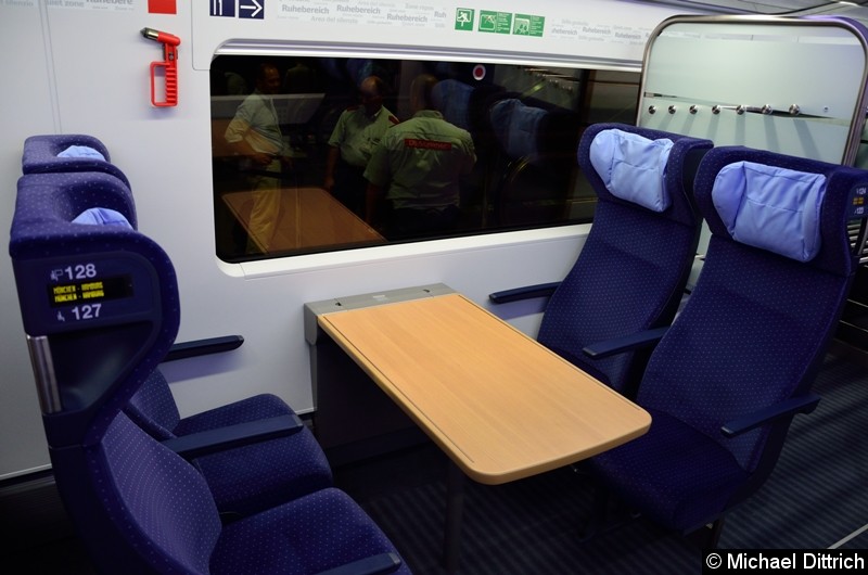 An diesem Tischplätzen sitzen zwei Personen neben einer Wand. Hinter den Sitzen gegenüber ist eine der vier Gepäckablagen des Zuges.